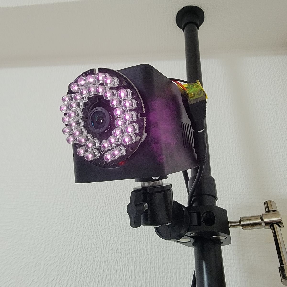 黒い3Dプリントのケースに入ったカメラ。前面で36個の850 nm LEDが光り、中央にM12レンズ。床から天井までの支柱に、クランプと小さな自由雲台で取り付けている